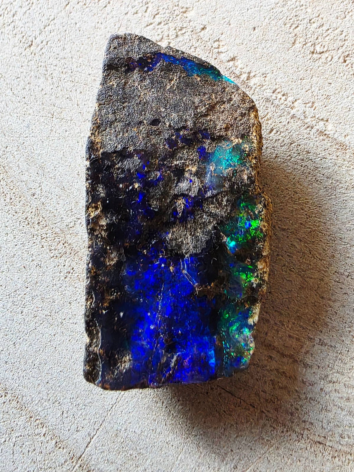 A rough raw opal stone showing vibrant blue, green, and purple iridescent play-of-colour against a textured light beige background.