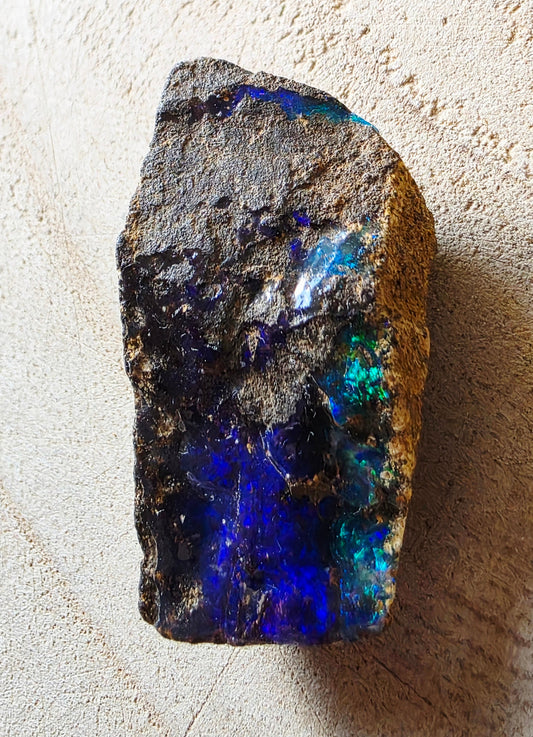 A rough natural opal stone with dark brown and black matrix, showing vibrant blue, green, and purple iridescent flashes, photographed on a textured beige surface.