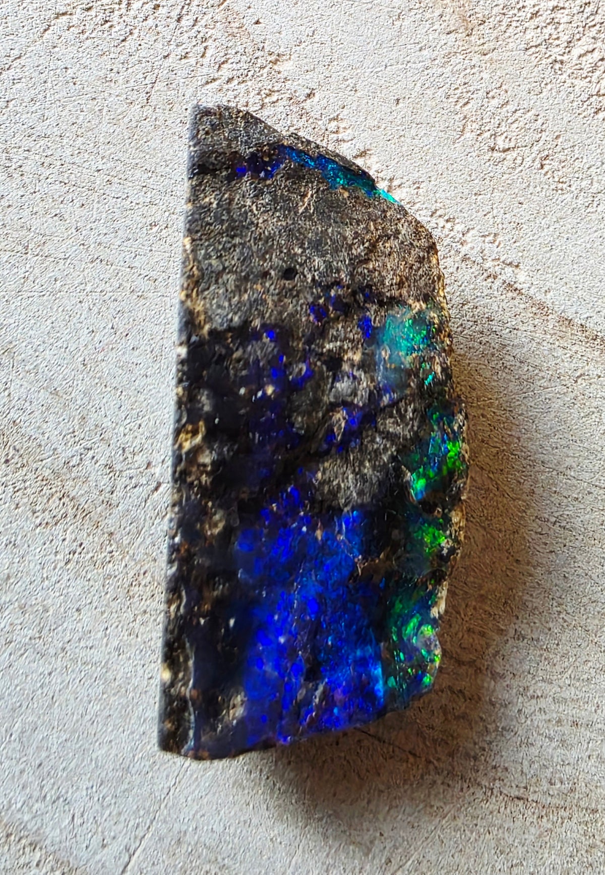 A raw opal stone with vibrant blue, green, and purple flashes on a dark, textured matrix, photographed on a light beige textured surface.