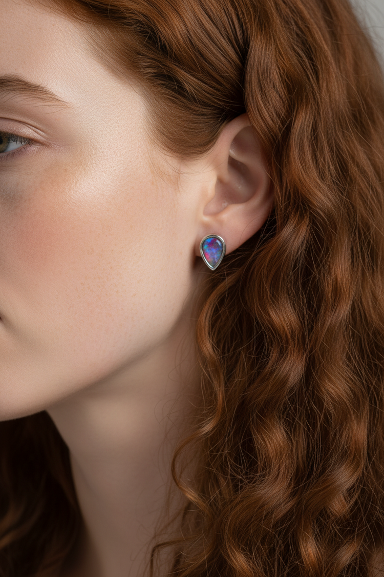 Close-up of a woman with wavy red hair wearing a small teardrop-shaped iridescent opal earring with a silver bezel setting.