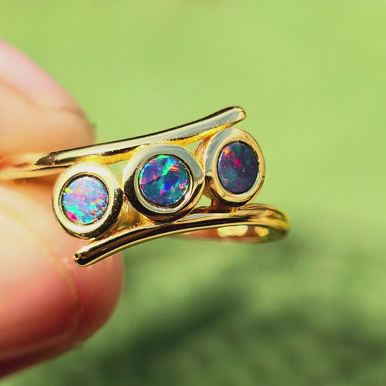 A gold ring featuring three round bezel-set iridescent opal stones, displayed on a textured beige background.