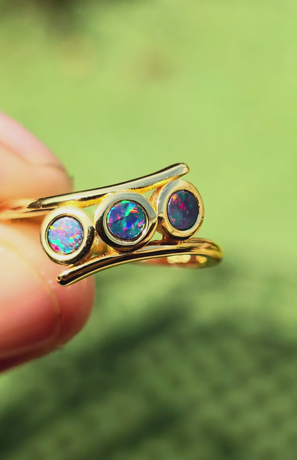 A gold ring featuring three round bezel-set iridescent opal stones, displayed on a textured beige background.
