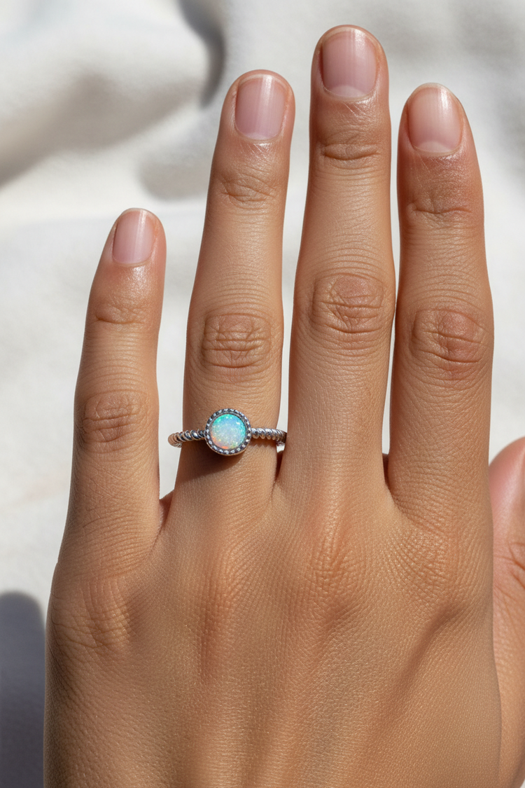 A close-up of a hand wearing a silver ring with a round iridescent blue-green opal-like centre stone.