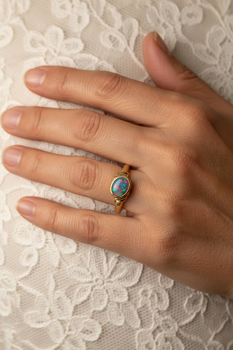 A gold ring with an oval multicolour opal gemstone, worn on a person's ring finger against a white lace background.