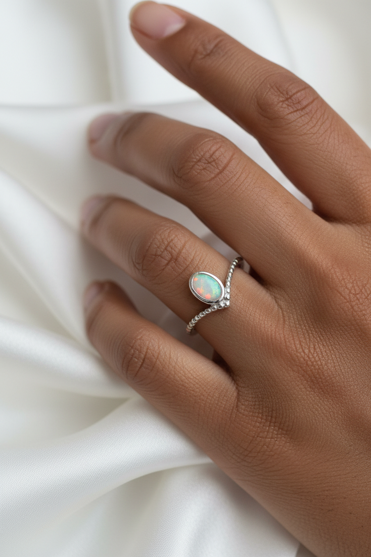 A close-up of a hand wearing a silver ring with an oval iridescent opal centre stone, resting against draped white fabric.